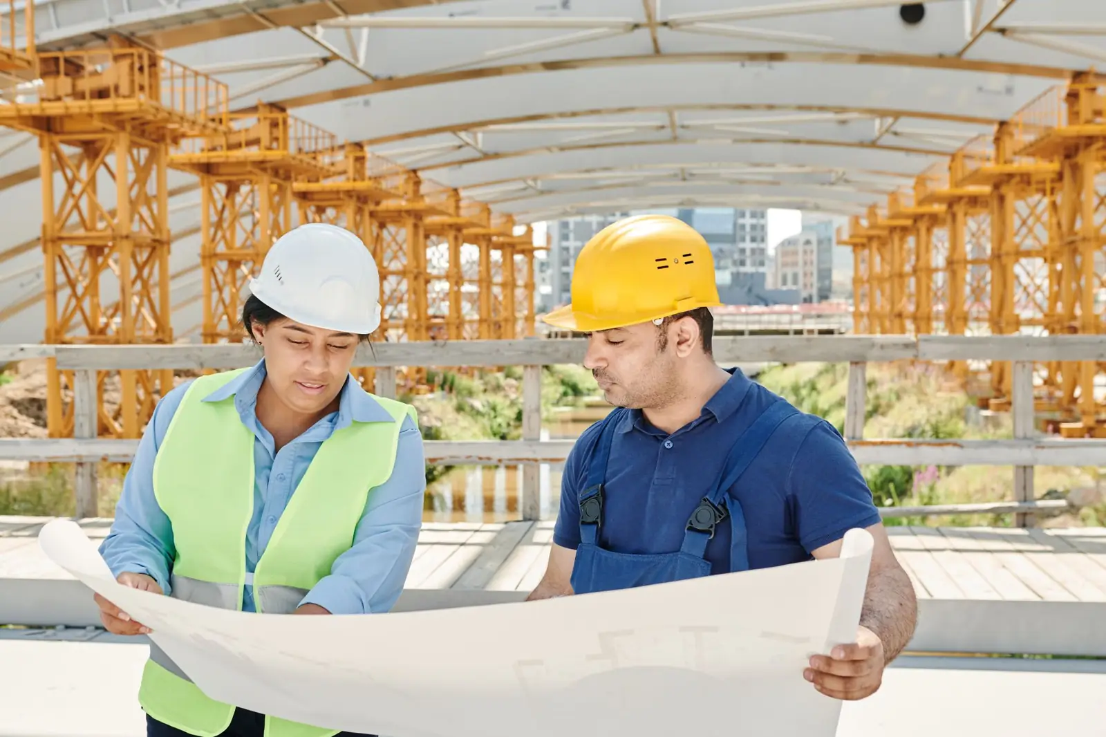 Two engineers in hard hats reading a drawing set on a steel-framed job site