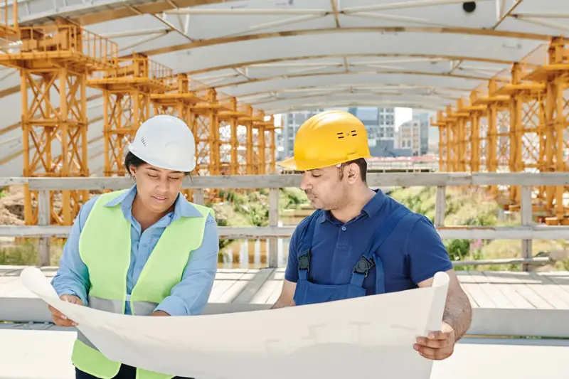 Two engineers in hard hats reading a drawing set on a steel-framed job site