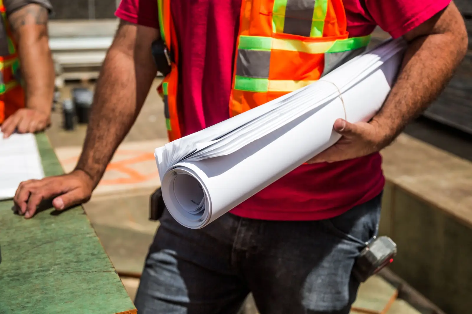 A site worker in a safety vest holding a roll of construction drawings
